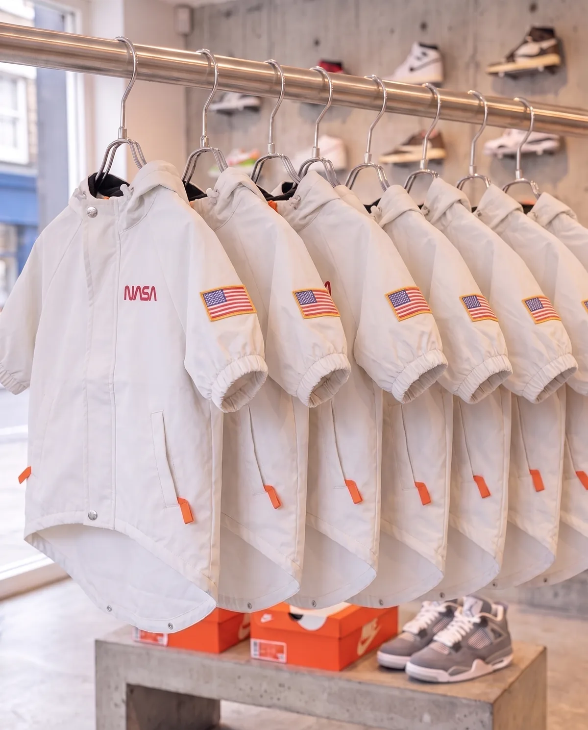 White NASA 3M Reflective Windbreakers on a chrome rack in a sneaker shop, front view with the small NASA chest logo and flag patch, flash off