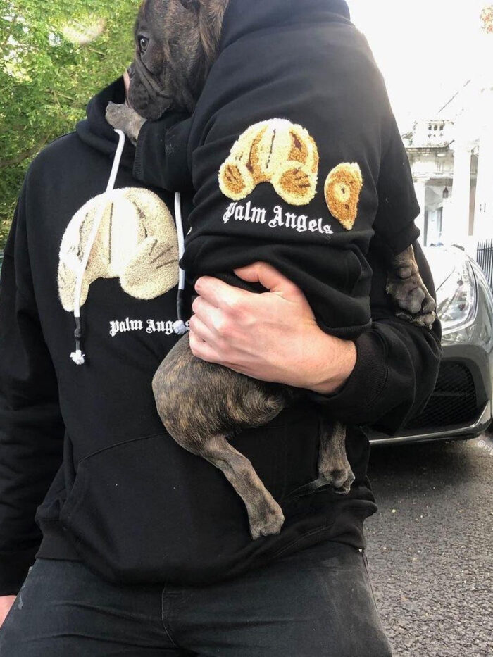 Person wearing black Palm Angels bear hoodie holding French bulldog in matching hoodie outdoors